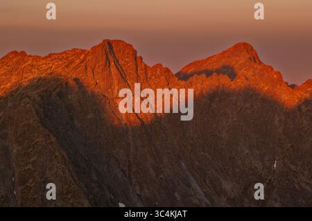 Vue aérienne des sommets montagneux incandescents capturant les teintes chaudes du soleil couchant, projetant des ombres dramatiques sur le terrain accidenté, Vysoké Tatry, région de Prešov, Slovaquie. Banque D'Images
