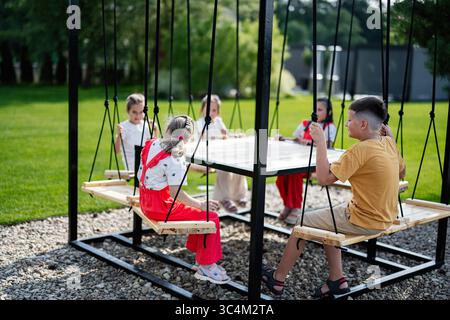 Groupe d'enfants assis sur des bancs oscillants autour d'une table par une journée ensoleillée Banque D'Images