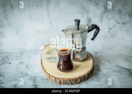Pot de moka, café noir et eau fraîche disposés sur un panneau rustique de tranche d'arbre contre béton léger, composition propre large prête pour les en-têtes de menu ou BA Banque D'Images