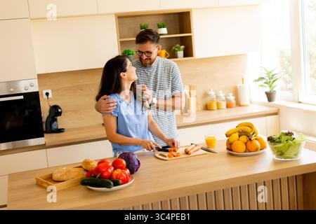 Un couple aime préparer un repas ensemble dans une cuisine lumineuse, entouré de fruits et légumes colorés, créant une atmosphère délicieuse remplie de w Banque D'Images