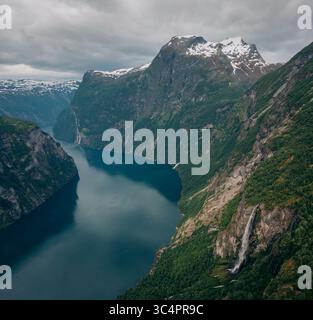 Vue aérienne des eaux sombres du Geirangerfjord serpentant entre d'imposantes falaises vertes et des cascades sous un ciel couvant, municipalité de Stranda, Banque D'Images