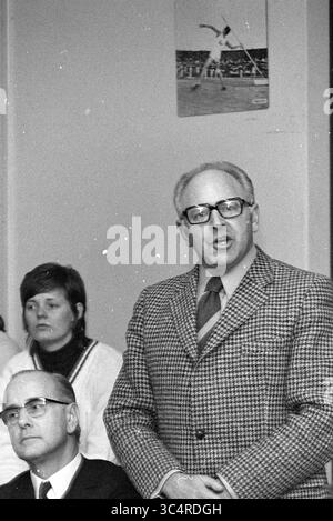 Actions chez CIO entraînement sportif chez Duinlust Whizgle News, Dutch Desk, pays-Bas, 1950-2000 Un homme portant des lunettes et un costume à motifs se tient devant, parlant passionnément, tandis que deux personnes derrière lui écoutent attentivement. L'une est une femme aux cheveux courts, et l'autre est un homme plus âgé en costume. Une affiche sportive est accrochée au mur en arrière-plan. Banque D'Images