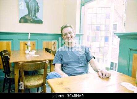 Frank Huissen, restaurant de Ark Haarlem, Haarlem, Nieuw Heiligland, pays-Bas, 28-10-1999 Whizgle News, Dutch Desk, pays-Bas, 1950-2000 Un homme détendu est assis à une table en bois dans un café confortable, souriant confortablement. L'intérieur lumineux présente des panneaux verts, avec une peinture d'une femme sur le mur à proximité. La lumière pénètre à travers une grande fenêtre, créant une atmosphère chaleureuse. Banque D'Images
