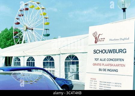 Construction Meerjazz, Hoofddorp, pays-Bas, 22-05-2002 Whizgle News, Dutch Desk, pays-Bas, 1950-2000 Une scène de festival dynamique avec une grande roue colorée surplombant une grande tente, avec un panneau annonçant des billets de concert gratuits et des représentations programmées. L'atmosphère suggère un événement animé, attirant les visiteurs à profiter de la musique et du divertissement. Banque D'Images