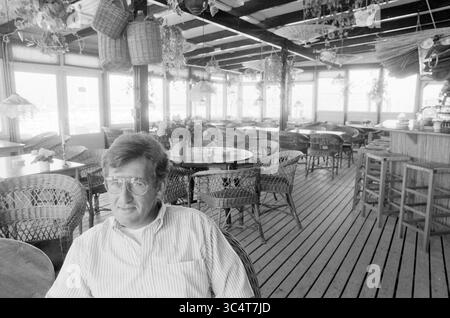 Propriétaire Freddy et Paul Meerendonk, Café, plage et plages, 31-05-1989 Whizgle News, Dutch Desk, pays-Bas, 1950-2000 Un homme est assis de façon décontractée dans un café spacieux et aéré avec du parquet et des meubles tissés, entouré de plantes et d'objets décoratifs suspendus au plafond. L'atmosphère est calme, créant une ambiance détendue dans l'établissement. Banque D'Images