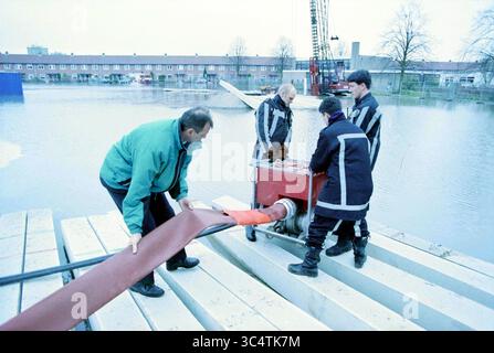 Rupture de tuyau d'eau R. Holkade, 09-03-1998 Whizgle News, Dutch Desk, pays-Bas, 1950-2000 Un groupe de quatre personnes travaille ensemble à la mise en place d'équipements près d'une zone inondée, avec des véhicules utilitaires et une grue de chantier en arrière-plan. Ils manipulent un gros tuyau et le connectent à une pompe, ce qui démontre des efforts de collaboration pour gérer l'eau. Banque D'Images