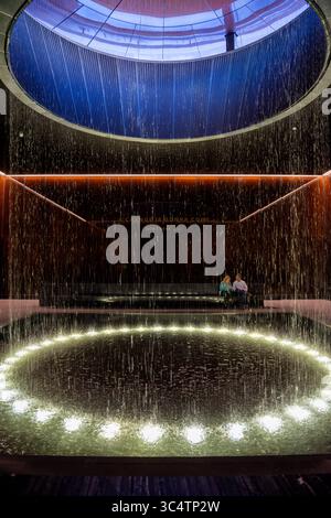 Musée national d'histoire et de culture afro-américaines fontaine de cour contemplative Washington DC | WASHINGTON D.C., États-Unis — Une vue verticale capture l'eau qui coule de l'oculus de la cour contemplative du Musée national d'histoire et de culture afro-américaines Smithsonian, encadrée par des murs de verre en cuivre-bronze. La cour occupe le niveau 0 du hall, fonctionnant comme un point de pause entre les galeries d'histoire du musée en dessous du niveau et les galeries culturelles au-dessus. Deux visiteurs sont assis sous une lyrique gravée de Sam Cooke, 'A change Is Gonna Come', l'une des nombreuses citations gravées dans t Banque D'Images