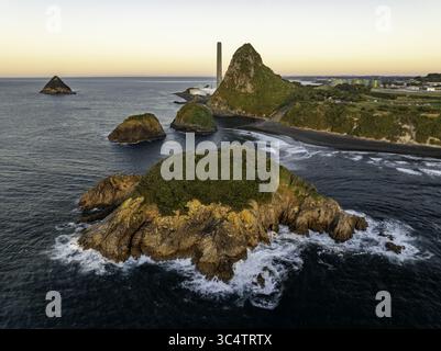 Vue aérienne des îles accidentées couvertes de mousse entourées de vagues à sommet blanc, contrastant avec la plage de sable sombre et le monolithe lointain, New Plymouth, région de Taranaki, Nouvelle-Zélande. Banque D'Images