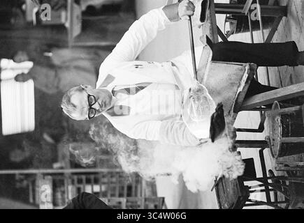 Visitez soufflage de verre, vitraux, soufflage de verre, verre en général, 14-06-1967 Whizgle News, Dutch Desk, pays-Bas, 1950-2000 Un artisan habillé d'une blouse de laboratoire manipule soigneusement le verre fondu, mettant en valeur l'art et la précision impliqués dans le soufflage du verre. De la vapeur et de la fumée s'élèvent autour de lui alors qu'il travaille sur un banc robuste, entouré de divers outils et équipements essentiels à son métier. Banque D'Images