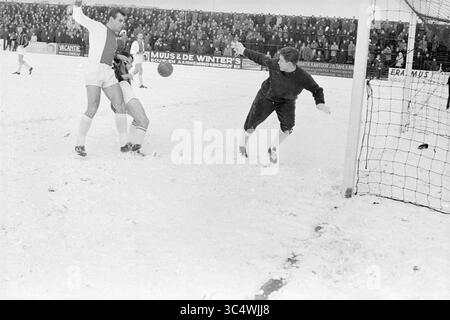 RCH - Ajax, Football, 17-02-1963 Whizgle News, Dutch Desk, pays-Bas, 1950-2000 Un moment dramatique pendant un match de football où un joueur glisse pour frapper le ballon alors que le gardien de but plonge pour faire une sauvegarde, le tout sur un terrain enneigé avec une foule regardant attentivement en arrière-plan. Banque D'Images