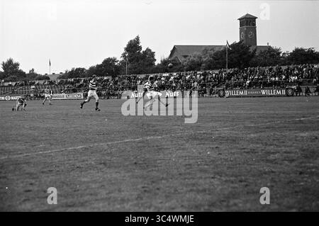 Football, Haarlem, pays-Bas, 00-00-1964 Whizgle News, Dutch Desk, pays-Bas, 1950-2000 Un joueur en pleine action passe le ballon lors d'un match de football intense, tandis que les spectateurs remplissent les gradins en arrière-plan, créant une atmosphère d'excitation et de compétition. Une tour d'horloge se profile au loin, ajoutant un sentiment de place à la scène. Banque D'Images