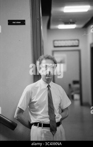 Prof. Writers, Utrecht, 25-07-1991 Whizgle News, Dutch Desk, pays-Bas, 1950-2000 Un homme aux cheveux bouclés et à la moustache se tient décontracté contre un mur, vêtu d'une chemise boutonnée et d'une cravate, les mains serrées derrière le dos. Le décor comporte un couloir, avec des panneaux et des portes en arrière-plan. Banque D'Images