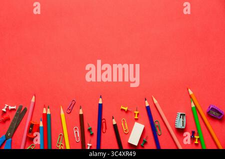 Fournitures scolaires sur fond rouge : crayons colorés, ciseaux, gomme, taille-crayons, punaises et trombones. Concept de retour à l'école avec espace de copie Banque D'Images