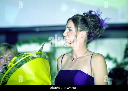 Mlle Hillegom 2002, Hillegom, 11-10-2002 Whizgle News, Dutch Desk, pays-Bas, 1950-2000 Une femme dans une élégante robe violette se tient gracieusement, souriante comme elle tient un bouquet vibrant de fleurs jaunes. Sa coiffure présente des boucles ludiques ornées d'éléments décoratifs, ajoutant à son apparence charmante. En arrière-plan, un autre individu semble parler, créant une atmosphère animée. Banque D'Images