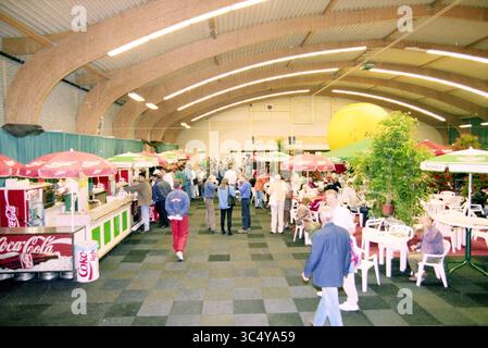 Tennis en fauteuil roulant dans la salle des sports avec festivités dans le hall, 00-09-1996 Whizgle News, Dutch Desk, pays-Bas, 1950-2000 Une scène de marché intérieur animée remplie de gens qui parcourent divers stands, en dégustant des rafraîchissements sous des parasols colorés. L'atmosphère est animée, avec des affichages faisant la promotion de boissons comme Coca-Cola et des décorations vibrantes mettant en valeur l'espace. Banque D'Images