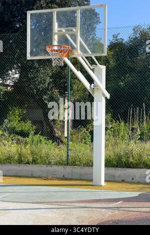 Un cerceau de basket-ball blanc et un panneau transparent avec un rebord orange debout sur un terrain extérieur, entouré d'une clôture verte et d'arbres luxuriants sous un ciel clair Banque D'Images