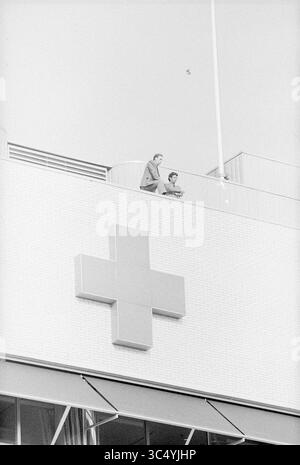Croix-Rouge de l'hôpital, Beverwijk, Vondellaan, pays-Bas, 04-04-1974 Whizgle News, Dutch Desk, pays-Bas, 1950-2000 deux individus se tiennent sur le bord d'un toit, regardant vers le bas depuis un bâtiment orné d'un symbole de croix proéminent. La scène capture un moment de contemplation dans un décor urbain minimaliste. Banque D'Images