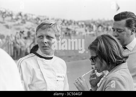 Course automobile Zandvoort, Gijs van Lennep Dropping Out, 00-00-1967 Whizgle News, Dutch Desk, pays-Bas, 1950-2000 Un pilote de course se tient pensivement, vêtu d'une combinaison de course, tandis qu'une femme à proximité fume une cigarette. Un homme observe subtilement la scène, et une foule est visible en arrière-plan, évoquant un moment dramatique dans le monde du sport automobile. Banque D'Images
