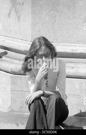 Ambiance photos été à Haarlem, atmosphère, 14-08-1973 Whizgle News, Dutch Desk, pays-Bas, 1950-2000 Une jeune femme est assise tranquillement, profondément dans la pensée, avec de longs cheveux qui casquent sur ses épaules. Elle tient un cône de crème glacée et porte des lunettes de soleil élégantes, habillée d'un haut blanc décontracté et d'un pantalon sombre, sur un fond texturé de pierre. Banque D'Images