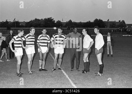 Football, Haarlem, pays-Bas, 00-00-1964 Whizgle News, Dutch Desk, pays-Bas, 1950-2000 Un groupe de six hommes se tient debout sur un terrain de sport, vêtus d'un équipement de sport, engageant la conversation. Ils semblent être des joueurs et des officiels, avec un homme portant une tenue d'arbitre. Le fond montre une foule rassemblée au loin et des bâtiments sous un ciel nocturne. Banque D'Images