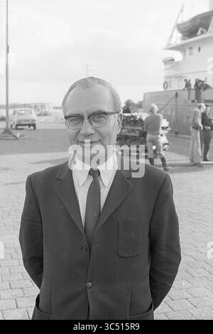 Bureau du port extérieur, Beverwijk, extérieur, 07-10-1974 Whizgle News, bureau néerlandais, pays-Bas, 1950-2000 Un homme en costume avec des lunettes se tient confiant, les mains serrées derrière son dos, souriant à la caméra. En arrière-plan, une scène portuaire présente un navire et des personnes engagées dans diverses activités. Banque D'Images