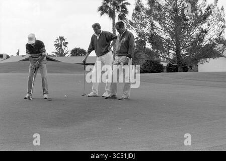 Visite des Gentlemen de Kinheim aux États-Unis, 09-11-1990 Whizgle News, Dutch Desk, pays-Bas, 1950-2000 trois hommes sont concentrés sur une balle de golf sur un putting green, l'un se préparant à tirer tandis que les autres observent de près. De grands palmiers et un fond résidentiel créent une atmosphère détendue typique d'un terrain de golf. Banque D'Images