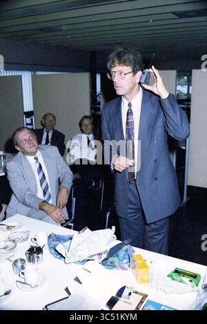 40e anniversaire A. Bot, fluor Daniel, 01-10-1991 Whizgle News, Dutch Desk, pays-Bas, 1950-2000 Un homme en costume se tient à une table, tenant un téléphone portable à son oreille tout en s'adressant à un public d'hommes assis, qui semblent engagés et curieux. La table est ornée de divers articles, y compris un cadeau froissé et une tasse de café. Banque D'Images