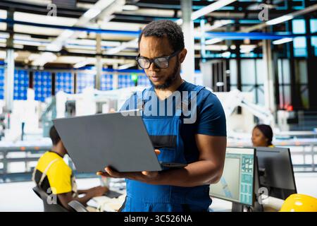 Ingénieur afro-américain ajuste les paramètres industriels pour la production d'énergie solaire, travaillant dans une usine de fabrication de haute technologie. Expert analysant les données techniques sur pc, finetuning. Banque D'Images