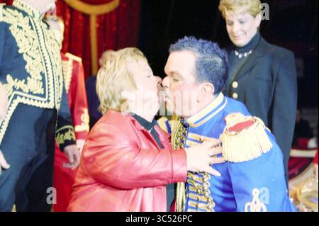 Décoration royale A. Olivier Circus Renz, 21-12-2002 Whizgle News, Dutch Desk, pays-Bas, 1950-2000 Une scène joviale mettant en scène un homme dans une veste de style militaire bleu vif partageant un baiser ludique avec une femme dans un manteau rouge saisissant, tous deux souriants. En arrière-plan, d'autres interprètes, vêtus de costumes élaborés, observent le moment joyeux. Banque D'Images