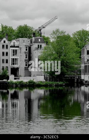 La vue depuis le pont Ezelsbrug à Amsterdam montre des maisons de canal et une grue reflétées dans l'eau calme sous un ciel sombre, la verdure adoucissant la scène. Banque D'Images