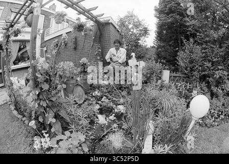 Femme posant dans (propre ?) Garden, 00-00-1993 Whizgle News, Dutch Desk, pays-Bas, 1950-2000 Une personne est assise avec soin dans un jardin luxuriant rempli de plantes, de fleurs et de verdure, entouré d'une arche en bois rustique ornée de décorations suspendues. Le cadre paisible dispose d'un mélange d'éléments de jardin, créant un espace extérieur harmonieux. Banque D'Images