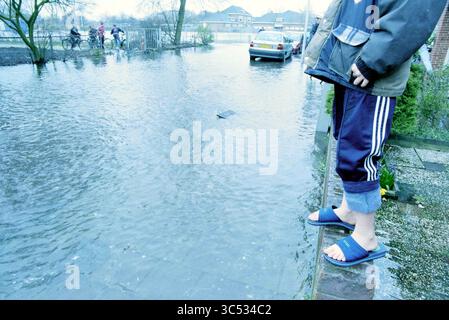 Burst Water pipe R. Holkade, 09-03-1998 Whizgle News, Dutch Desk, pays-Bas, 1950-2000 Une personne se tient au bord d'une rue inondée, portant une tenue décontractée et des pantoufles, tandis que les cyclistes à proximité naviguent sur le chemin inondé. En arrière-plan, des wagons partiellement submergés sont visibles, indiquant l'étendue de l'inondation. Banque D'Images