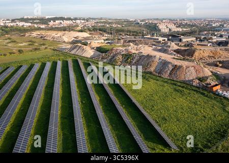 Des rangées de panneaux solaires couvrent un champ vert vibrant adjacent à un chantier de construction. Ciel dégagé et toile de fond suburbaine soulignent le contraste entre nature et développement. Banque D'Images