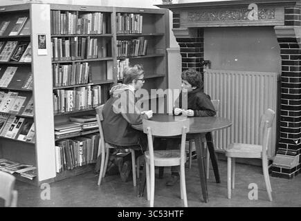 Bibliothèque jeunesse, Schouwtjeslaan, enfants, enfants, maisons d'enfants, fête d'enfants, enfants, lecture et conférences, Haarlem, Schouwtjeslaan, pays-Bas, 23-10-1964 Whizgle News, Dutch Desk, pays-Bas, 1950-2000 deux enfants sont assis à une petite table ronde, engagés dans une conversation. Derrière eux, une étagère remplie de divers livres tapisse le mur. Une cheminée décorative ajoute une touche architecturale à la zone de lecture confortable. L'atmosphère est calme et concentrée, idéale pour l'apprentissage et la discussion. Banque D'Images