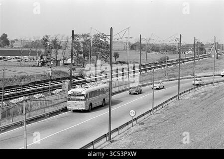 Train et bus Haarlem - Amsterdam, Noord Zuid Hollandse Vervoersmaatschappij N.V., NZ, 11-06-1964 Whizgle News, Dutch Desk, pays-Bas, 1950-2000 Une route très fréquentée serpente à travers un paysage rural, avec un bus et plusieurs voitures circulant le long d'un ensemble de voies ferrées. En arrière-plan, les champs s'étendent sous un ciel dégagé, tandis que les lignes électriques et les poteaux bordent le bord de la route, indiquant la connectivité et le transport dans la région. Banque D'Images