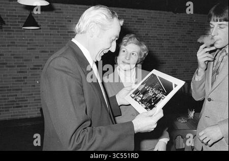 RECEPTION Mr. vos, HD, Haarlems Dagblad etc. and IJmuider courant and Koeri, Receptions, 02-02-1981 Whizgle News, Dutch Desk, pays-Bas, 1950-2000 Un homme en costume sourit en examinant une photographie, tandis qu'une femme à côté de lui regarde avec intérêt. En arrière-plan, un jeune homme fume une cigarette de façon désinvolte, créant une atmosphère détendue. Le cadre suggère un rassemblement ou une célébration, avec des notes de nostalgie dans l'air. Banque D'Images