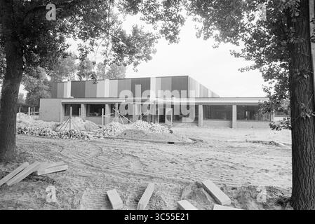 INR. Et ext. Salle de sport IJmuiden Brunostraat, salles de sport, IJmuiden, pays-Bas, 09-07-1979 Whizgle News, Dutch Desk, pays-Bas, 1950-2000 Un bâtiment moderne en construction, avec une combinaison de panneaux colorés et de grandes fenêtres, entouré de piles de matériaux de construction et d'un paysage sablonneux. Des arbres imposants encadrent la scène, ajoutant une touche de nature au site en développement. Banque D'Images