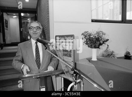 Ouverture du bâtiment HD Holding, 06-05-1991 Whizgle News, Dutch Desk, pays-Bas, 1950-2000 Un homme en costume se dresse sur un podium avec un microphone, souriant en parlant. Derrière lui, un journal est affiché à côté d'un vase décoratif de fleurs, et un petit arrangement de plantes ajoute au décor. Le fond révèle un intérieur moderne avec un aperçu des escaliers et des portes vitrées. Banque D'Images