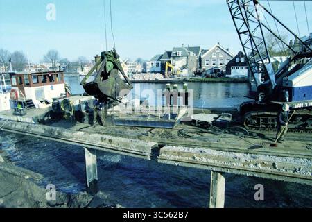 Activités Spaarne, Haarlem, pays-Bas, 12-02-1998 Whizgle News, Dutch Desk, pays-Bas, 1950-2000 Un chantier de construction le long d'une rive est équipé de machinerie lourde, dont une grue et une barge, activement engagées dans des travaux d'excavation. On voit les travailleurs gérer l'équipement alors qu'ils préparent la zone pour le développement. En arrière-plan, une charmante ville riveraine ajoute une toile de fond pittoresque à l'activité animée. Banque D'Images