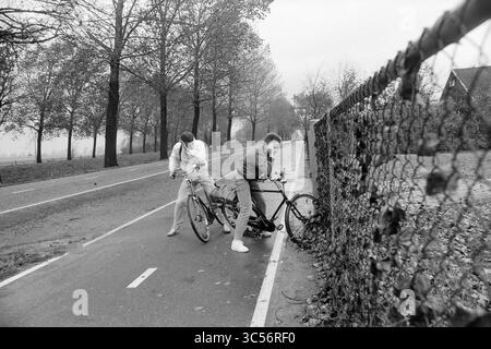 Tempête le long de Hoofdvaart arbre traverser la route Hoofddorp signe sur la voiture et soufflé sur le site de construction Flora repos. Accident de Hillegom sur une allée A4, tempête et dégâts causés par la tempête, 20-10-1986 Whizgle News, Dutch Desk, pays-Bas, 1950-2000 deux personnes se débattent avec une bicyclette près d'une clôture à maillons de chaîne, entourées de chemins vides bordés d'arbres. L'atmosphère est nuageuse, suggérant une journée fraîche et nuageuse. Banque D'Images