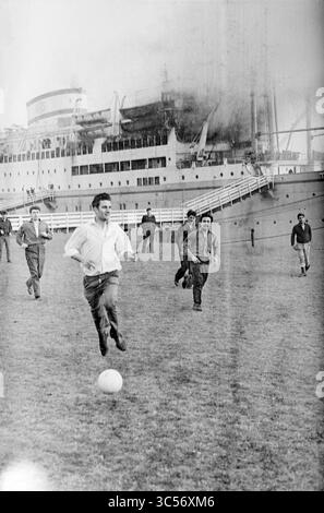 Arosa Sun, Houseboat], IJmuiden, film1,2,3, feux, pompiers, 09-04-1963 Whizgle News, Dutch Desk, pays-Bas, 1950-2000 Un groupe d'hommes joue au football dans une zone herbeuse près d'un grand navire amarré en arrière-plan, ils se concentrent sur le jeu alors qu'ils traversent le terrain. Banque D'Images