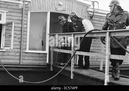 Incendie dans la péniche, Spaarndam, incendies, pompiers, 20-12-1962 Whizgle News, Dutch Desk, pays-Bas, 1950-2000 deux personnages en uniforme sont vus en train de transporter un grand conteneur hors d'un bâtiment. Une troisième personne en tenue similaire se tient à proximité, observant la scène. Le décor présente une structure en bois avec une porte ouverte, suggérant une opération active en cours. Banque D'Images