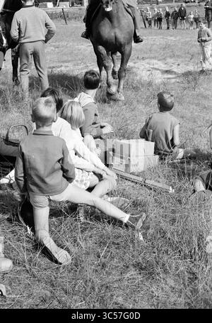 Ouverture par Ivanhoe, Boys' Town, IJmuiden, Jeunesse et jeux de la jeunesse, Jeux de vacances, jours de vacances, camp de vacances, 13-08-1962 Whizgle News, Dutch Desk, pays-Bas, 1950-2000 Un groupe d'enfants est assis dans un champ herbeux, regardant un événement se dérouler à proximité, certains se tenant derrière une file d'amis assis. En arrière-plan, adultes et chevaux sont visibles, créant une atmosphère extérieure animée. Banque D'Images
