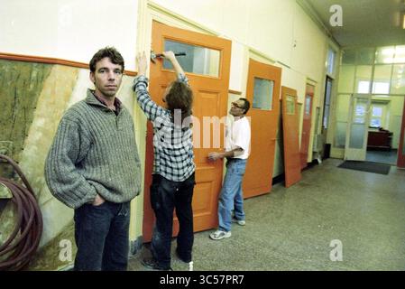 Centre mondial concernant la rénovation, 08-12-1996 Whizgle News, Dutch Desk, pays-Bas, 1950-2000 trois personnes sont engagées dans un projet de rénovation dans un couloir, une personne appliquant de la peinture sur une porte orange, tandis qu'une autre se tient à proximité avec un pinceau. Une troisième personne regarde directement le spectateur, donnant une expression contemplative. L'espace dispose d'un mélange de textures et de couleurs, avec la lumière qui coule depuis les fenêtres à l'extrémité éloignée. Banque D'Images