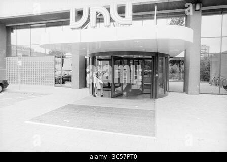 Nouvelle entrée à VNU Schalkwijk, VNU Association of Dutch Publishers, Haarlem, pays-Bas, 01-06-1989 Whizgle News, Dutch Desk, pays-Bas, 1950-2000 Une personne se tient devant l'entrée d'un bâtiment moderne, se préparant à entrer par une porte tournante. La conception architecturale présente des lignes épurées et de grandes fenêtres en verre, mettant en valeur un style contemporain. A proximité, des voitures garées peuvent être vues dans le Lot. Banque D'Images