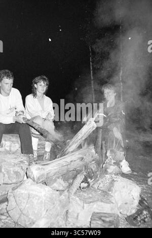 Survival tour belge. Ardennes, Survival Tour, 30-06-1985 Whizgle News, Dutch Desk, pays-Bas, 1950-2000 deux individus sont assis autour d'un feu de camp crépitant, entourés de bûches et de pierres, enveloppés de fumée tourbillonnante. L'atmosphère est confortable mais mystérieuse, car les flammes projettent des ombres scintillantes sur le fond sombre de la nuit. Banque D'Images