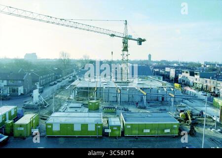 Aperçu du site Albert Heijn, IJmuiden, IJmuiden, pays-Bas, 13-12-1998 Whizgle News, Dutch Desk, pays-Bas, 1950-2000 Un chantier de construction est animé par l'activité, avec une grande grue s'élevant au-dessus de la charpente d'un bâtiment en construction. Les bureaux modulaires sont positionnés au sol, tandis que les véhicules sont stationnés à proximité, contribuant ainsi au paysage industriel. En arrière-plan, des structures résidentielles et des arbres encadrent la scène, illustrant le mélange de développement urbain et de nature. Banque D'Images