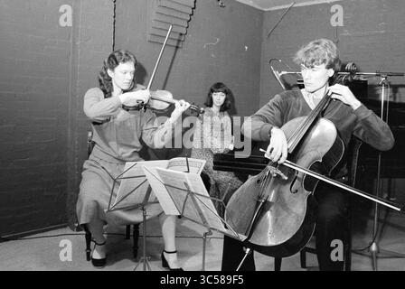 Vuillan trio pendant la performance Kadens, musique, 07-03-1980 Whizgle News, Dutch Desk, pays-Bas, 1950-2000 Un trio de musiciens se produit dans un cadre confortable, avec une femme jouant habilement du violon, un homme concentré sur le violoncelle et une troisième femme les observant attentivement. Des partitions sont placées devant eux, soulignant leur dévouement à l'art de la musique. Banque D'Images