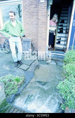 Rupture de la conduite d'eau R. Holkade, 09-03-1998 Whizgle News, Dutch Desk, pays-Bas, 1950-2000 Un homme se tient tranquillement devant une résidence, regardant vers le bas l'eau qui coule le long du chemin, tandis qu'une femme regarde hors de la porte, observant la situation. Une bicyclette s'appuie contre le mur à proximité, et la scène transmet un sentiment d'inquiétude au sujet de l'écoulement inattendu de l'eau. Banque D'Images