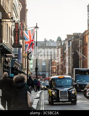 Un taxi londonien noir classique traverse Soho devant les drapeaux de l'Union Jack et les piétons, capturant une scène de rue britannique par excellence. Banque D'Images